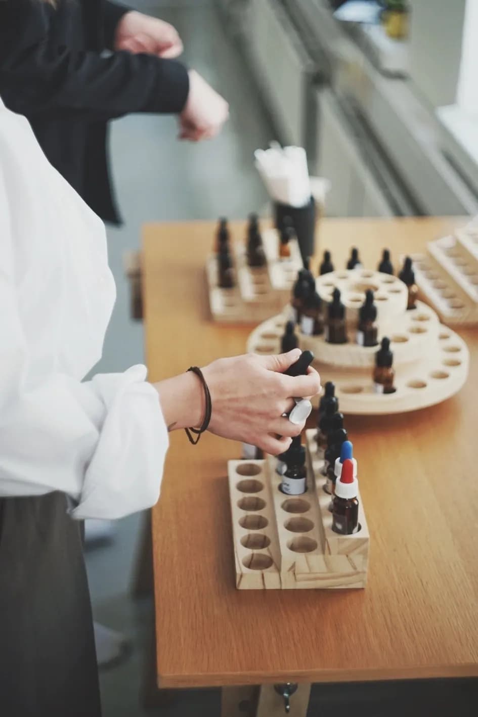A workshop guest lifting a dropper bottle from a wooden rack of perfume materials